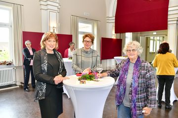 Frau Grodzki, Frau Behrens und Frau Dr. Karras stehen um einen Stehtisch mit Blumen darauf und lächeln in die Kamera. Im Hintergrund sind die roten Stoffbahnen als Dekoration in der Aula und weitere Personen zu sehen)