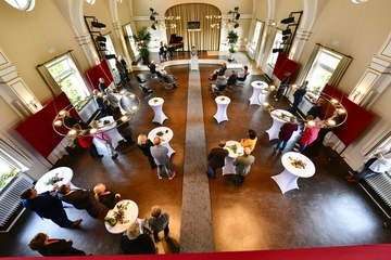 Die Aula des LBZB von oben (Die Aula des LBZB ist von oben zu sehen. Im (Hintergrund steht eine Person am Rednerpult, davor sitzen einige Personen auf Stühlen. Im Vordergrund stehen Personen an Stehtischen)