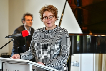 Frau Ministerin Behrens (Frau Ministerin Behrens steht an einem hellen Rednerpult. Im Hintergrund ist der Pianist Herr Dr. Hribar sitzend am Flügel zu sehen)