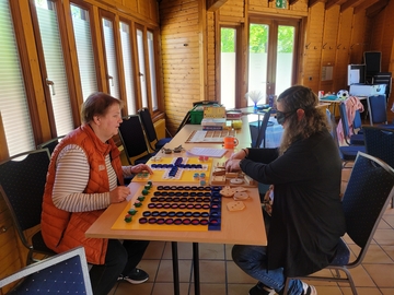 Zwei Teilnehmerinnen sitzen sich an einer Tischreihe gegenüber und spielen Gesellschaftsspiele. Eine Teilnehmerin spielt unter der Augenbinde, die andere gibt ihr Hilfestellung.