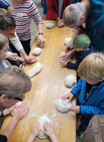 Kinder und Erwachsene stehen an einem Tisch und formen Brotlaibe aus einem Stück Teig.
