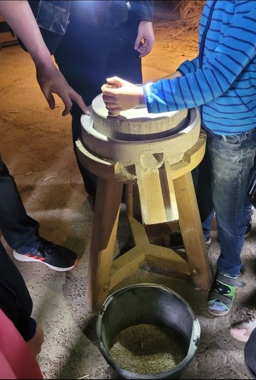 Ein Junge dreht einen Mahlstein - unter dem Stein sind Getreidekörner. Das gemahlene Korn rutscht auf einer kleinen Holzbahn in einen Metalleimer.