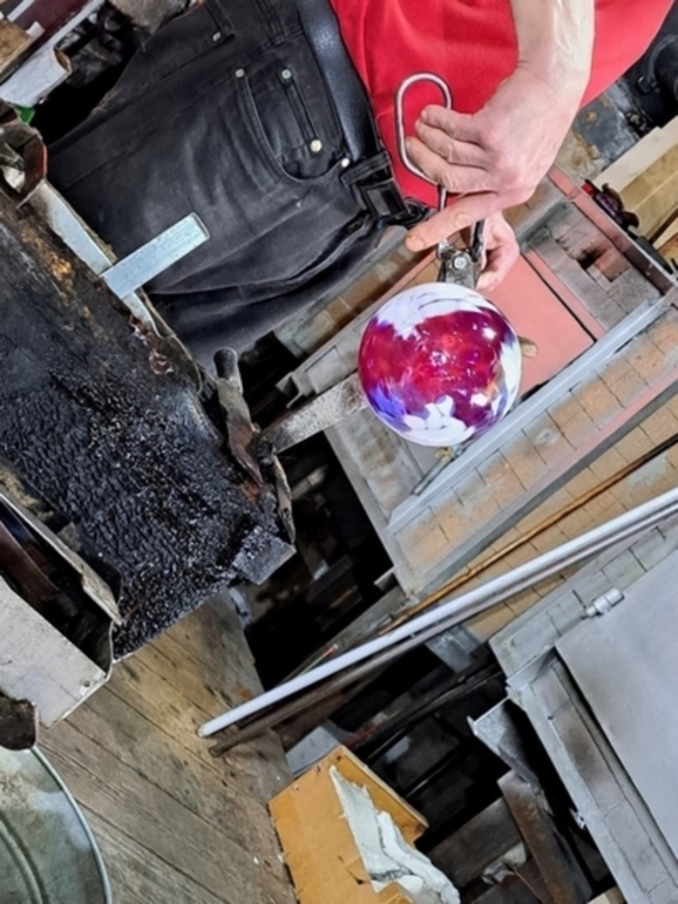 Zu Besuch in einer Glasbläserei. Jemand hat gerade eine bunte Glaskugel gedreht und aufgepustet, nun wird sie abgeschnitten.