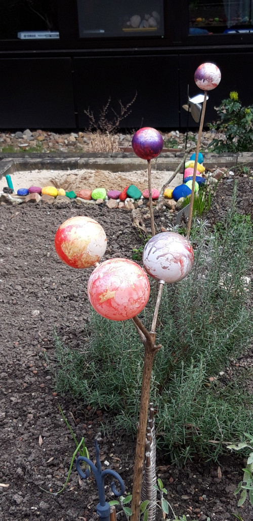 Blick über das Hochbeet Im Vordergrund Gartenkugeln auf Stöcken und Rosmarin im Beet, im Hintergrund unser selbstgebauter Sandkasten, begrenzt mit bemalten Steinen.