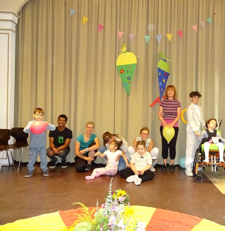 Schüler:innen sitzen, hocken und stehen mit einer Lehrerin auf einer Bühne.