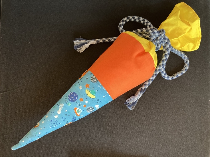 Auf einem schwarzen Untergrund liegt eine Schultüte. Sie besteht an der unteren Spitze aus einem Stoff mit Weltraum-Motiven. Daran schließt sich ein orangefarbener Stoff an. Den Abschluss bildet ein gelber Stoff. Die Öffnung der Schultüte ist mit ein