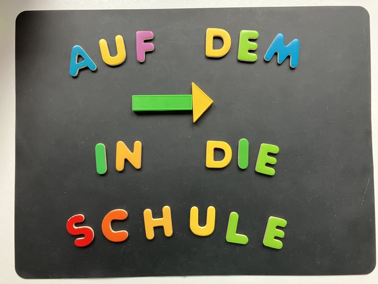 Auf einer schwarzen Unterlage ist mit bunten Großbuchstaben und Formen in vier Zeilen folgender Schriftzug gelegt: 1. Zeile: „AUF DEM“ 2. Zeile: Ein Pfeil nach rechts aus einem Rechteck und einem Dreieck. 3. Zeile: „IN DIE“ 4. Zeile: „SCHULE