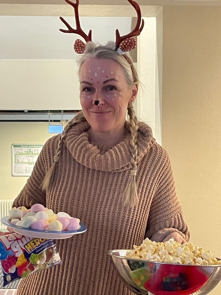 Frau Börner, als schön geschminktes und mit Hörnern auf dem Kopf verkleidetes Rentier, bringt eine große Schale mit Popcorn, einen Teller mit Marshmallow und eine Tüte Lachgummi.