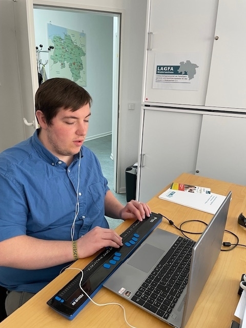 Ein junger Mann sitzt vor einem Laptop und vor seiner Tastatur liegt eine Braillezeile-Tastatur.