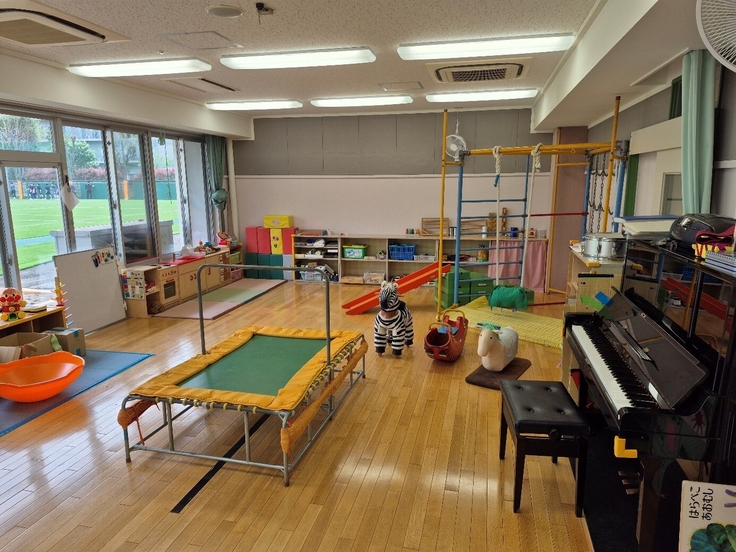 Auf dem Bild ist ein Raum des Kindergartens zu sehen. In dem Raum stehen in der Mitte ein kleines Trampolin und Spielzeug, rechts ein Klavier und in den Ecken Schränke.