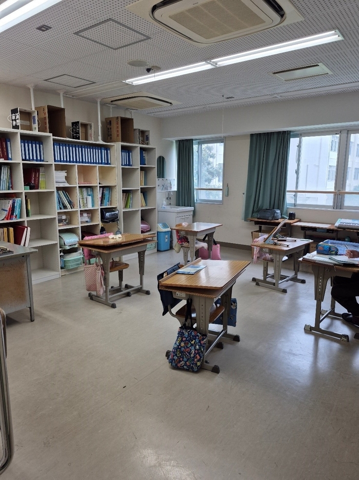 Auf dem Bild ist ein Klassenzimmer zu sehen. Es stehen mehrere Tische mit Stühlen jeweils einzeln im Raum. Im hinteren Bereich des Raumes steht ein großes Regal mit Büchern an der Wand.