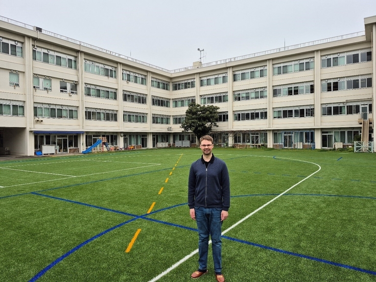 Auf dem Bild ist ein Mann zu sehen. Er steht auf einem Kunstrasen Sportplatz. Auf dem Sportfeld sind verschiedene Linien gemalt. Im Hintergrund ist das Schulgebäude zu erkennen.