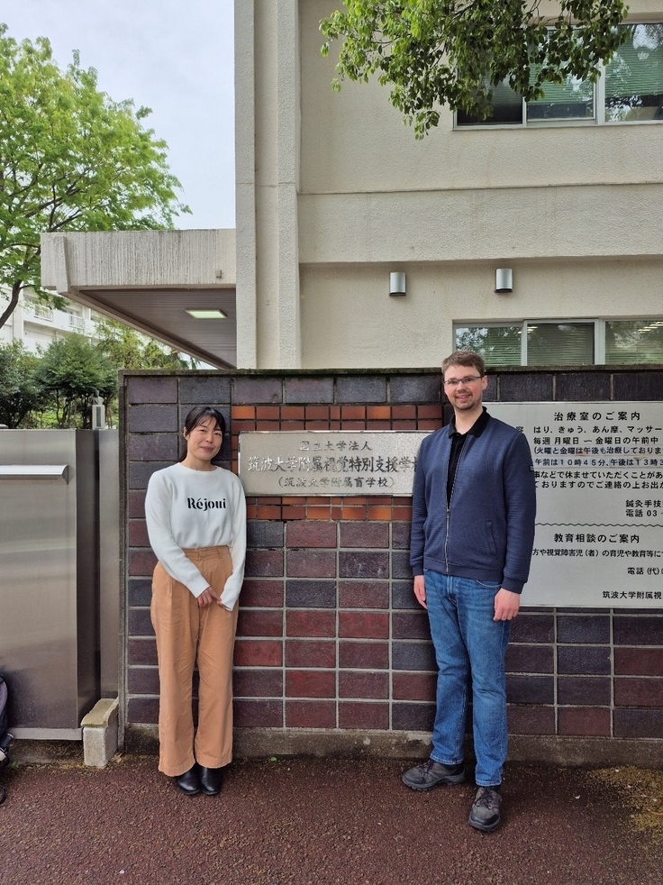 Auf dem Bild sind eine Frau und ein Mann zu erkennen. Beide stehen vor einer Mauer. An der Mauer ist ein Schild mit dem Namen der Schule befestigt. Das Schild ist in japanischer Schrift. Im Hintergrund ist das Schulgebäude zu erkennen.