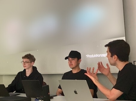 Drei Männer sitzen vor einer Leinwand den Zuhörenden zugewandt und beschreiben Ideen zu einer Hilfsmittelentwicklung.