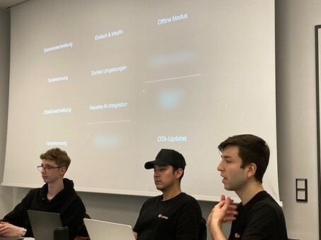 Drei Männer sitzen vor einer Leinwand den Zuhörenden zugewandt und beschreiben einzelne Elemente des Hilfsmittels, die im Hintergrund auf der Leinwand in Stichworten dargestellt sind.