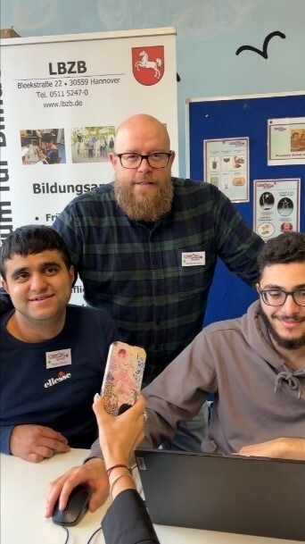 Ein Ausbilder und zwei Auszubildende warten am Messestand der LaserTec auf Kunden.