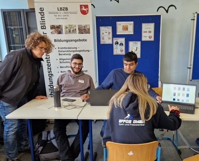 Eine Sozialarbeiterin und zwei Auszubildende führen ein Verkaufsgespräch mit einer Kundin, die sich gerade am Laptop einen Überblick über unser Sortiment verschafft.