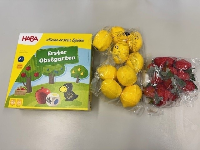 Auf dem Foto ist das Gesellschaftsspiel der Firma HABA „Erster Obstgarten“ für Kinder ab 2 Jahren abgebildet. Daneben sind 9 künstliche Zitronen und 12 künstliche Erdbeeren in Cellophantüten zu sehen.
