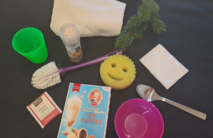 Auf einem schwarzen Untergrund befinden sich ein grüner Becher, ein weißes Handtuch, eine Gewürzmühle, ein grüner Tannenzweig, ein weißes Verpackungspapier, ein silberner Esslöffel, ein pinkes Schälchen, ein gelber Schwamm, eine lila Spülbürste