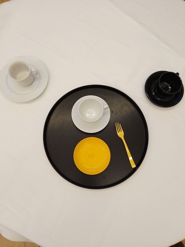 Auf dem Foto ist eine weiße Tischdecke zu sehen, auf der im Zentrum ein schwarzes Tablet platziert ist. Auf dem Tablet ist ein gelber Teller und eine gelbe Gabel zu sehen. Ebenso eine weiße Tasse, die auf einem weißen Unterteller steht. Am linken Bild