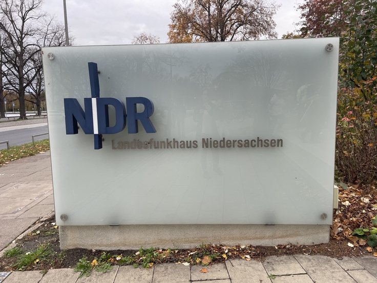 Auf einem großes weißes Hinweisschild vor dem Gebäude des NDRs steht in blauen Großbuchstaben "NDR", darunter in schwarzer, etwas kleinerer Schrift "Landesfunkhaus Niedersachsen"