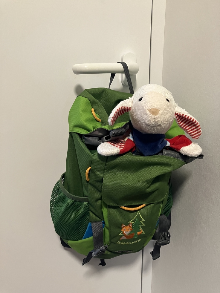 An einer weißen Türklinke hängt ein dunkelgrüner Rucksack. Aus der oberen Öffnung schauen der Kopf und die Vorderbeine eines Kuscheltier-Schafs heraus.