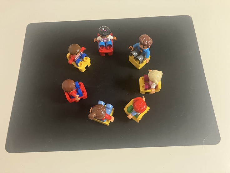 Auf dem Foto sind auf einer schwarzen Unterlage sieben Stühle aus Duplo-Steinen zu einem Sitzkreis angeordnet. Auf den Stühlen sitzen fünf kindliche Duplo-Männchen und zwei erwachsene Duplo-Männchen.