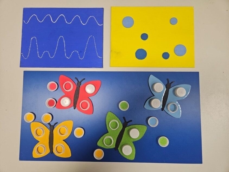 Auf dem Foto sind 3 Aufgabentafeln zu sehen. Auf einer blaue Aufgabentafel sind weiße tastbare Punkte zu sehen, die in Wellenform über die Tafel laufen. Auf einer gelben Tafel sind verschieden große und unterschiedlich tastbare Punkte in zwei Blautön