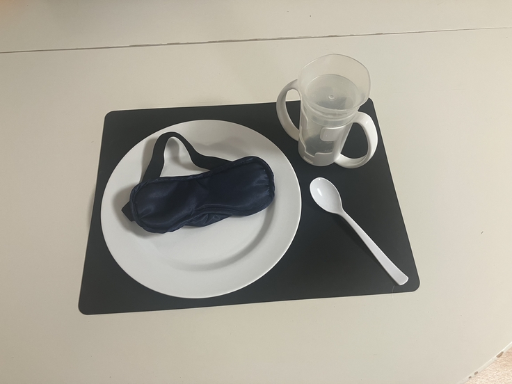 Auf einem schwarzen Set liegt links ein weißer Teller mit einer Schlafmaske darauf. Oben rechts steht ein Trinklernbecher mit Handgriffen. Unten rechts liegt ein weißer Löffel.