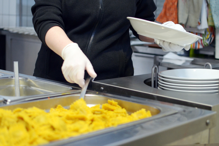 Eine Küchenkraft nimmt aus einer großen Warmhalteschale Raviolis heraus und legt sie auf einen Teller. Natürlich trägt sie zum Schutz Handschuhe.