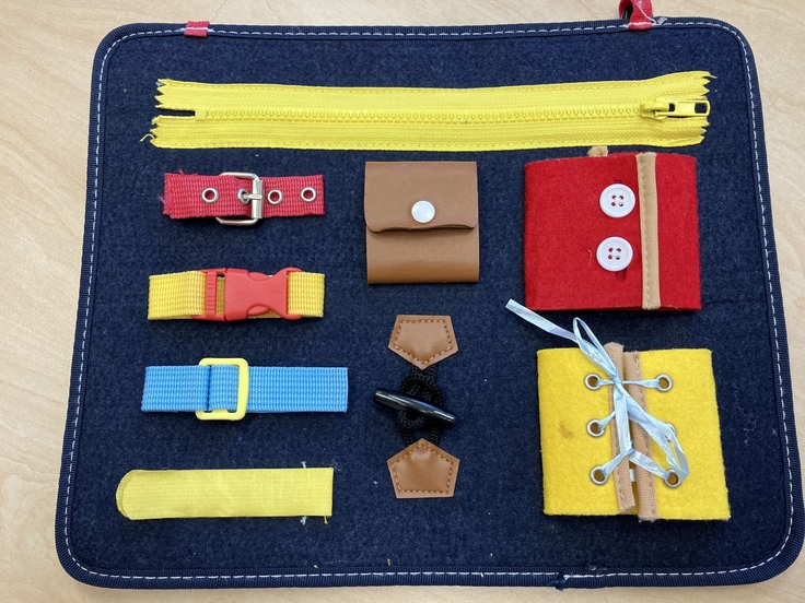 Das Bild zeigt ein Fördermaterial mit dem man lernen kann, Verschlüsse an Kleidung zu öff-nen und zu schließen. Auf einer Filzmatte sind Verschlüsse mit Knöpfen, Reißverschluss, Steckschnallen, Klett und zum Zubinden aufgenäht.