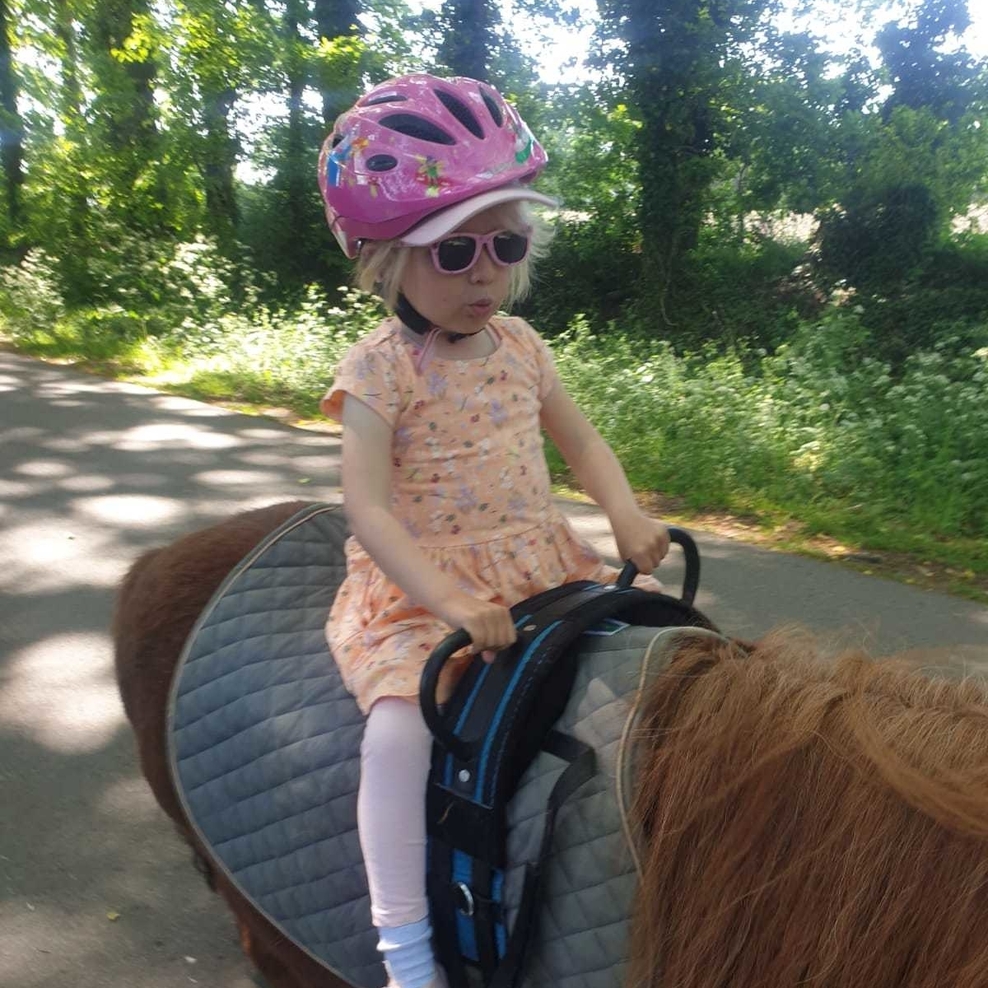 Auf dem Foto ist ein kleines Mädchen abgebildet, das auf einem braunen Pferd sitzt. Sie befindet sich auf einem Feldweg. Das Mädchen ist mit einem geblümten Kleid, einer langen Leggins, Socken und Sandalen bekleidet.