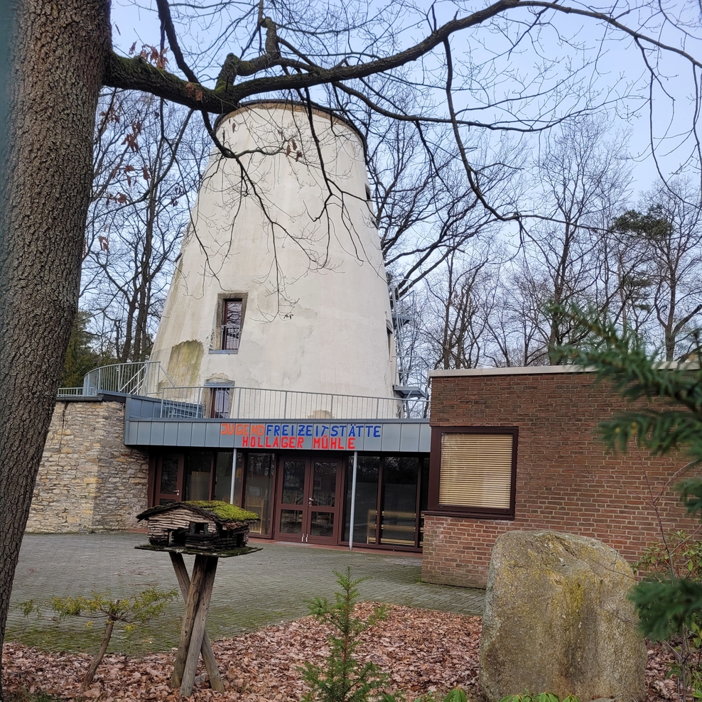 Auf dem Foto ist die Vorderansicht der Jugend- Freizeitstätte Hollager Mühle zu sehen. Im Vordergrund ist links ein Vogelhäuschen und rechts ein großer Findling zu sehen.
