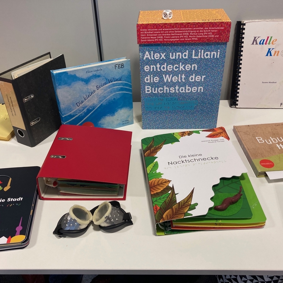 Das Bild zeigt einen Tisch mit verschiedenen Fühlbüchern für Kinder sowie einer Simulationsbrille.