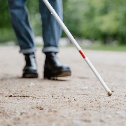 Auf einem sandigen Weg geht eine Person entlang. Nur die Beine und Schuhe sind auf dem Bild. Die Person hält einen Langstock. Im Hintergrund sind verschwommen Bäume und zwei weitere Personen auf einem Rasenstück zu erkennen.