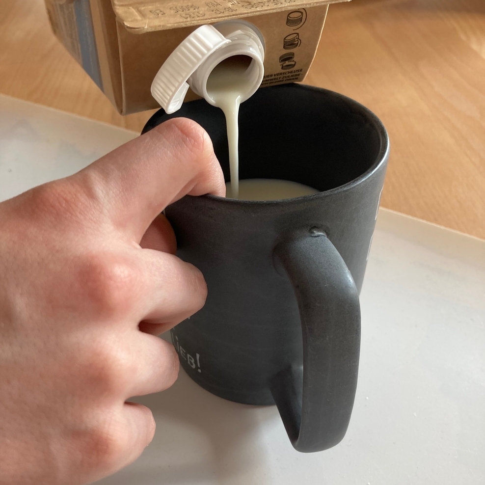 Auf einem weißen Set steht eine schwarze Tasse. Eine Hand greift mit dem Zeigefinger von oben am Rand in die Tasse. In die Tasse wird Milch eingegossen.