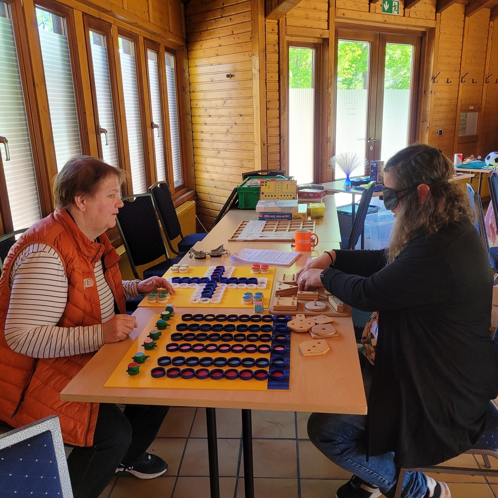 Zwei Teilnehmerinnen sitzen sich an einer Tischreihe gegenüber und spielen Gesellschaftsspiele. Eine Teilnehmerin spielt unter der Augenbinde, die andere gibt ihr Hilfestellung.