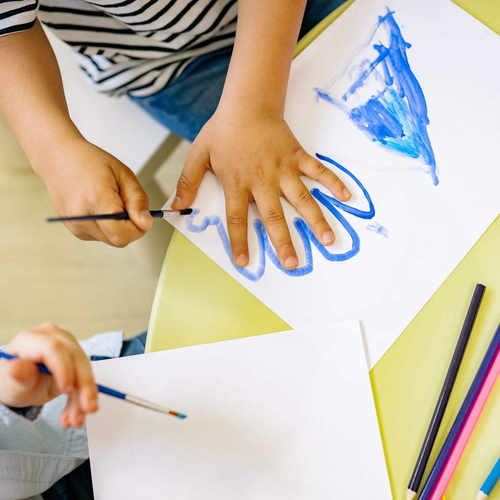 Auf einer Tischplatte sind drei Kinderhände beim Malen mit Pinseln und Wasserfarbe zu sehen. Es liegen Papierbögen auf dem Tisch. Eine Hand malt gerade mit blauer Farbe um die andere Hand herum.