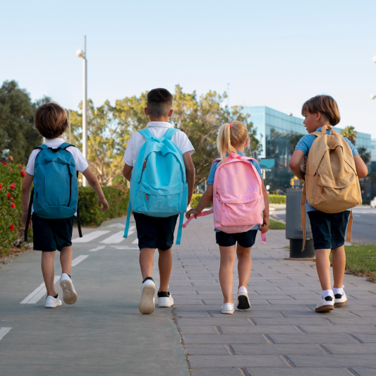 vier Kinder mit Schulrucksäcken gehen zusammen auf einem Bürgersteig in die Schule. Sie tragen sommerliche Kleidung.