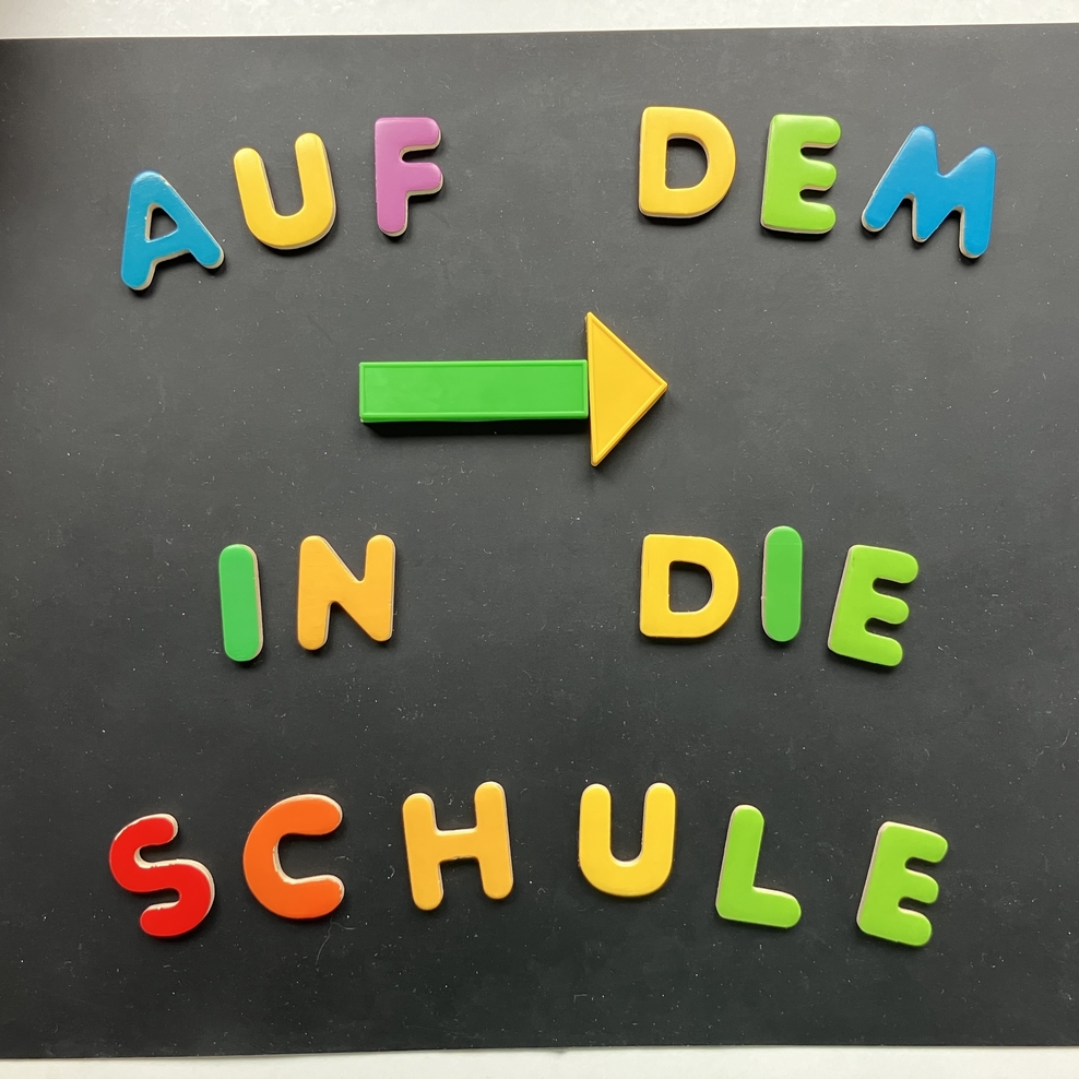 Auf einer schwarzen Unterlage ist mit bunten Großbuchstaben und Formen in vier Zeilen folgender Schriftzug gelegt: 1. Zeile: „AUF DEM“ 2. Zeile: Ein Pfeil nach rechts aus einem Rechteck und einem Dreieck. 3. Zeile: „IN DIE“ 4. Zeile: „SCHULE