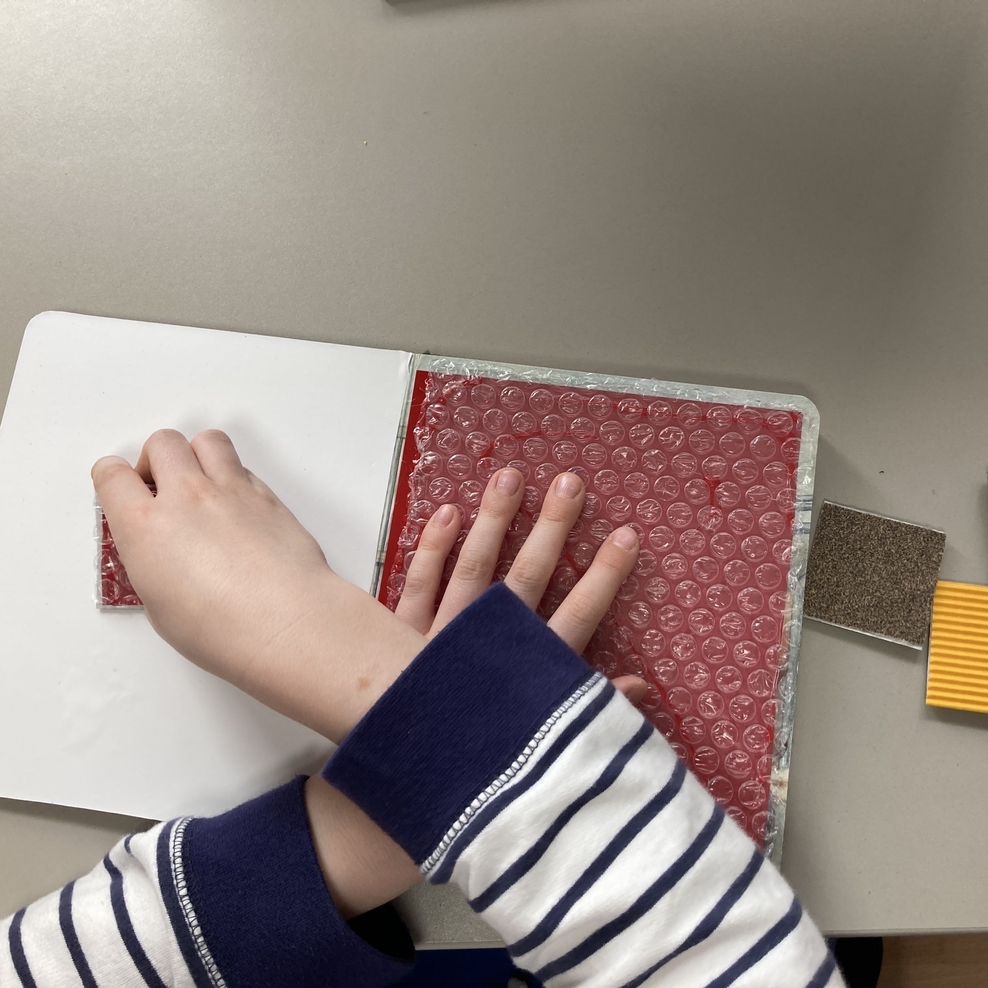 Auf einer Arbeitsfläche liegt ein aufgeschlagenes Buch. Auf der rechten Buchseite befindet sich auf rotem Untergrund Luftpolsterfolie. Rechts neben dem Buch liegen drei kleine quadra-tische Kärtchen mit verschiedenen Tastqualitäten. Eine linke Kinderh