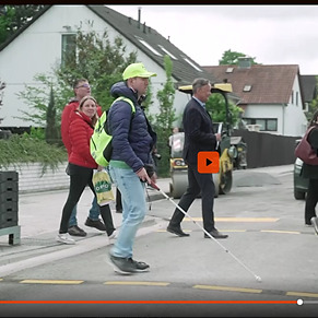 Mehrere Personen sehende und eine blinde Person überqueren eine Straße mit einer Baustelle.