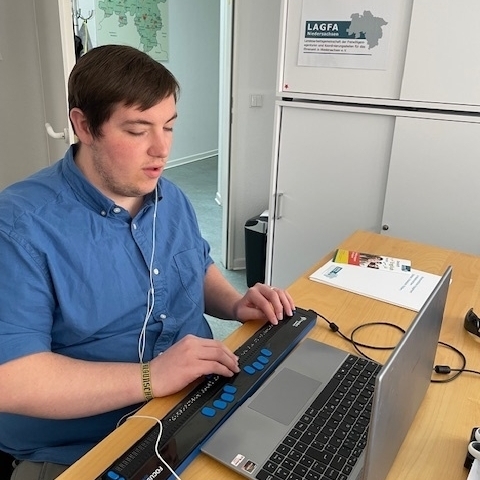 Ein junger Mann sitzt vor einem Laptop und vor seiner Tastatur liegt eine Braillezeile-Tastatur.