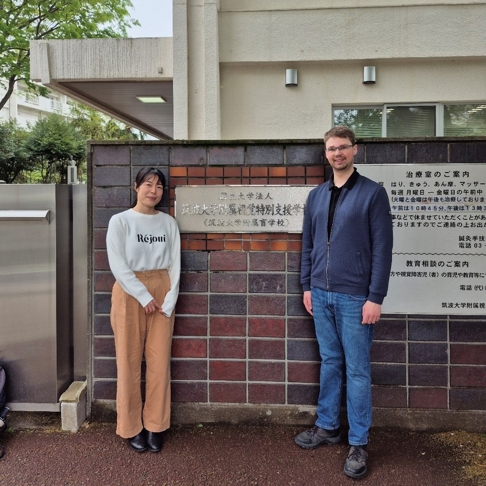 Auf dem Bild sind eine Frau und ein Mann zu erkennen. Beide stehen vor einer Mauer. An der Mauer ist ein Schild mit dem Namen der Schule befestigt. Das Schild ist in japanischer Schrift. Im Hintergrund ist das Schulgebäude zu erkennen.