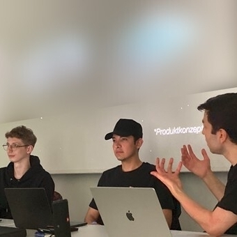 Drei Männer sitzen vor einer Leinwand den Zuhörenden zugewandt und beschreiben Ideen zu einer Hilfsmittelentwicklung.