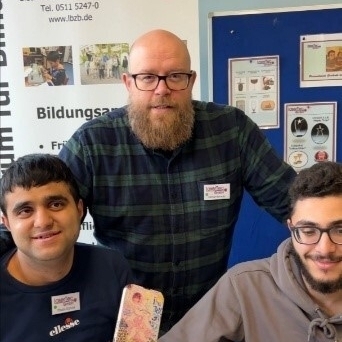 Ein Ausbilder und zwei Auszubildende warten am Messestand der LaserTec auf Kunden.