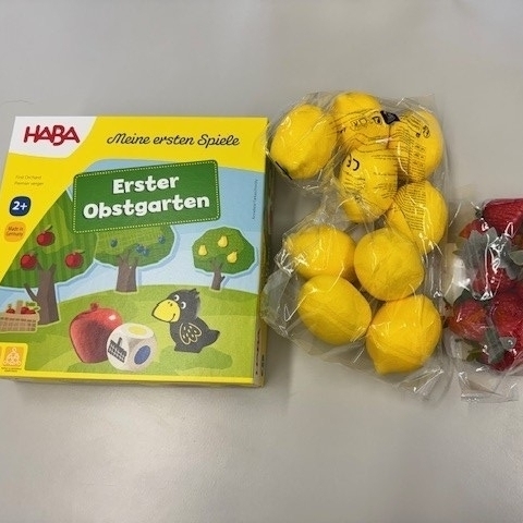 Auf dem Foto ist das Gesellschaftsspiel der Firma HABA „Erster Obstgarten“ für Kinder ab 2 Jahren abgebildet. Daneben sind 9 künstliche Zitronen und 12 künstliche Erdbeeren in Cellophantüten zu sehen.