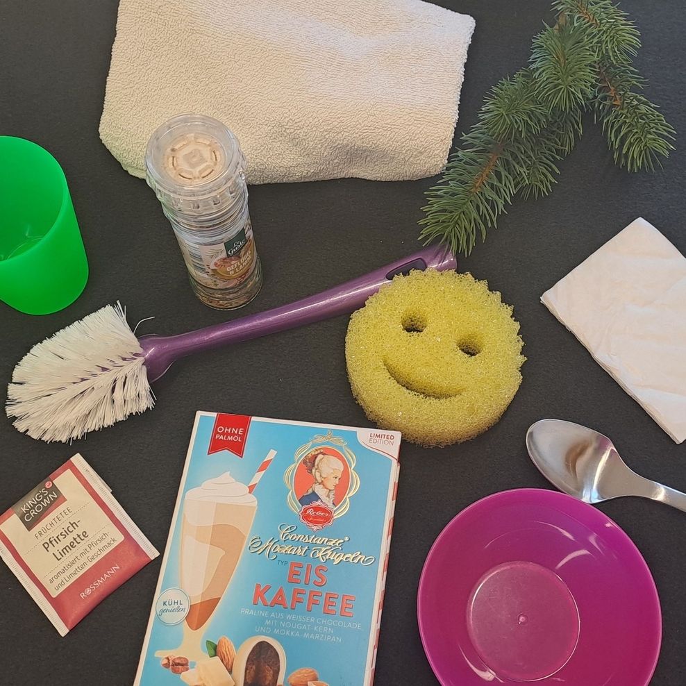 Auf einem schwarzen Untergrund befinden sich ein grüner Becher, ein weißes Handtuch, eine Gewürzmühle, ein grüner Tannenzweig, ein weißes Verpackungspapier, ein silberner Esslöffel, ein pinkes Schälchen, ein gelber Schwamm, eine lila Spülbürste