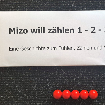 Auf einem schwarzen Untergrund liegen dicht beieinander 5 rote Kugeln. Darüber liegt ein weißes Schild. Darauf steht: Mizo will zählen 1-2-3-4-5. Eine Geschichte zum Fühlen, Zählen und Vorlesen lassen.