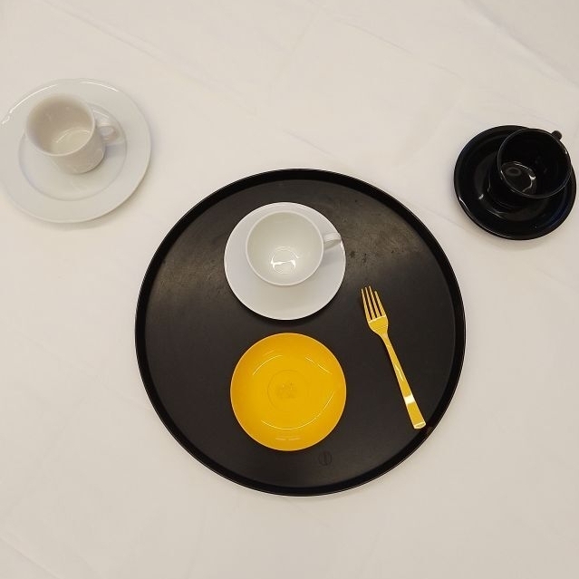 Auf dem Foto ist eine weiße Tischdecke zu sehen, auf der im Zentrum ein schwarzes Tablet platziert ist. Auf dem Tablet ist ein gelber Teller und eine gelbe Gabel zu sehen. Ebenso eine weiße Tasse, die auf einem weißen Unterteller steht. Am linken Bild